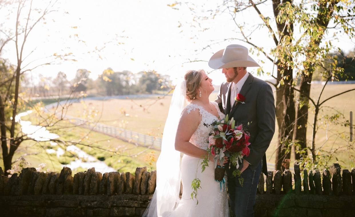 Bride and groom in a cowboy hat along the stone wall overlooking the pasture
