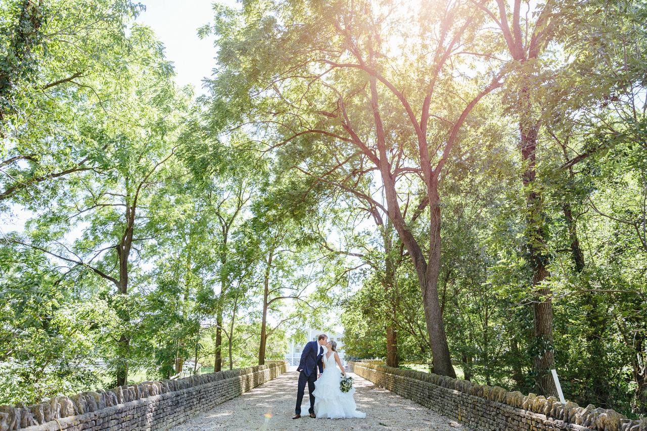 Bride and groom kissing on the stone bridge beneath sunlit trees